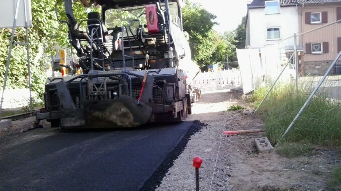 User comments im Vordergrund frischer Asphalt. Asphaltmaschine steht vor der Kreuzung mit der Straße Im Defdahl.