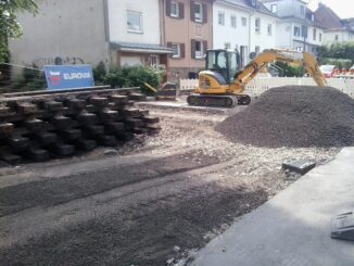 Links liegen gestapelte Gleise, rechts ein Haufen Schotter. Im Hintergrund stehen Häuser und davor ein Bagger.