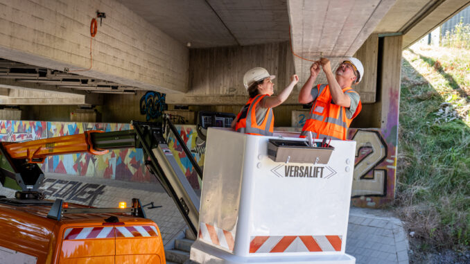 Mitarbeiter des Tiefbauamtes bringen einen der Sensoren an. Oben im Bild ist die Unterseite der Brücke. In der Bildmitte ist der Korb eines Hubwagens. Links im Korb steht eine Frau, rechts ein Mann. Sie montieren über Kopf die Sensoren. Links im Bild sieht man ein Teil des Montagewagens.
